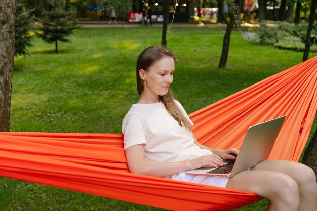 한 젊은 여성 이 나무 들 사이 에 있는 오렌지색 하마크 에 앉아 노트북 에 타이핑 하고 있다