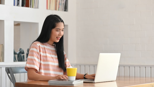 The young woman is using a computer laptop while sitting at the wooden working desk