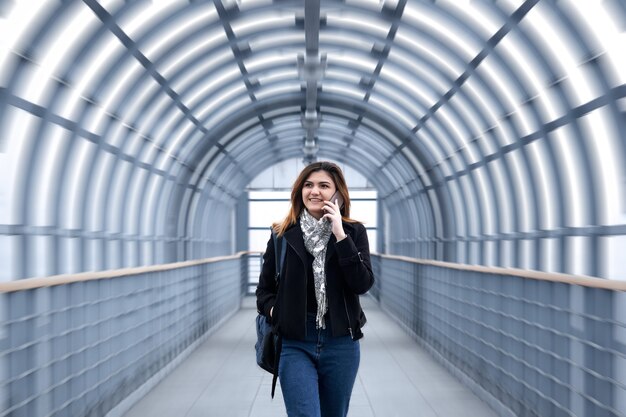 Young woman is walking rapidly along a covered pedestrian overpass, talking on the phone and smiling