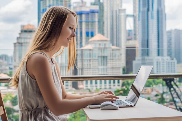 Young woman is working on a laptop on her balcony overlooking the skyscrapers Freelancer remote work work from home