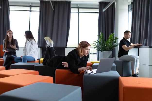 A young woman is working with a laptop lying on ottomans in the office