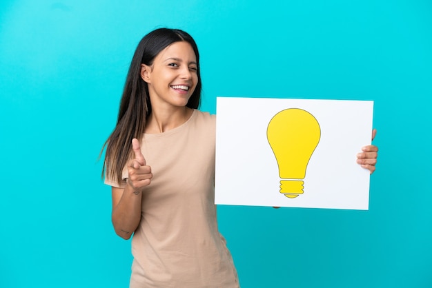 Young woman over isolated background holding a placard with bulb icon and pointing to the front