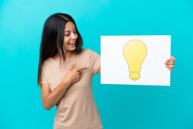 Young woman over isolated background holding a placard with bulb icon and pointing it