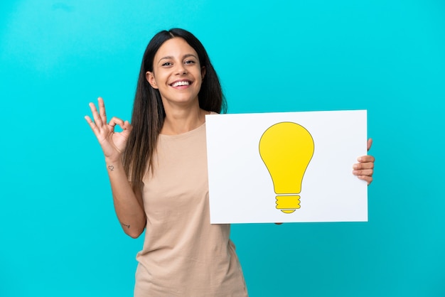 Young woman over isolated background holding a placard with bulb icon with ok sign