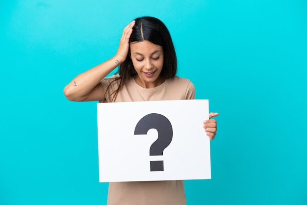 Young woman over isolated background holding a placard with question mark symbol with surprised expression