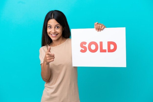 Young woman over isolated background holding a placard with text SOLD and pointing to the front