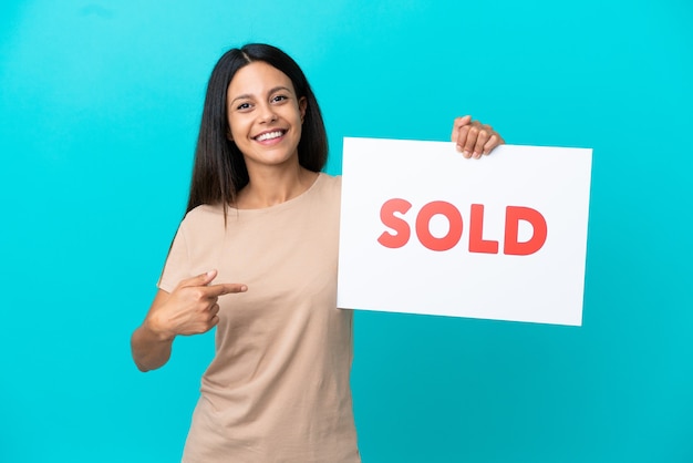 Young woman over isolated background holding a placard with text SOLD and  pointing it