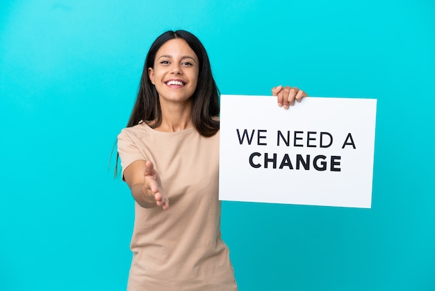 Young woman over isolated background holding a placard with text We Need a Change making a deal