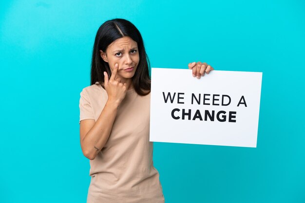 Young woman over isolated background holding a placard with text We Need a Change and showing something