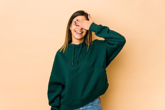 Young woman isolated on beige covers eyes with hands, smiles broadly waiting for a surprise.