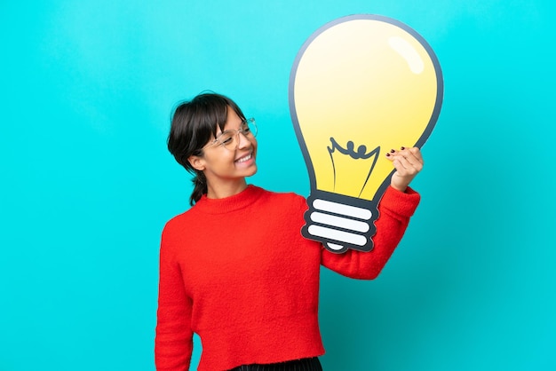 Young woman isolated on blue background holding a bulb icon with happy expression