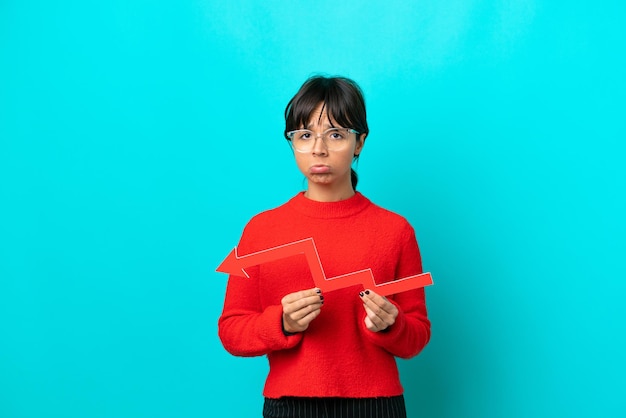 Young woman isolated on blue background holding a downward arrow and with sad expression