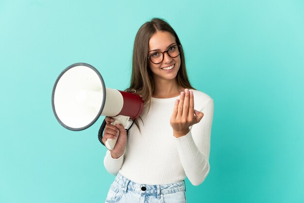 Foto giovane donna su sfondo blu isolato che tiene un megafono e che invita a venire con la mano