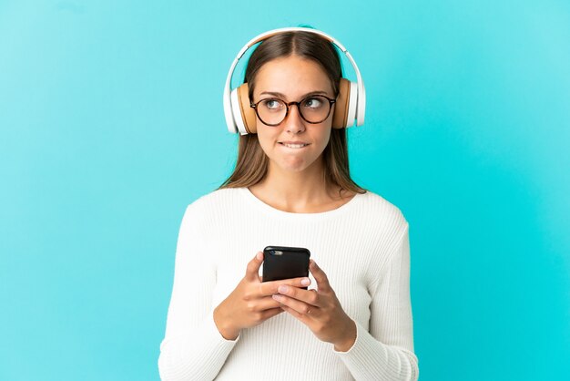Молодая женщина на изолированном синем фоне слушает музыку с мобильным телефоном и думает