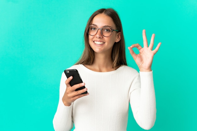 Young woman over isolated blue background using mobile phone and doing OK sign