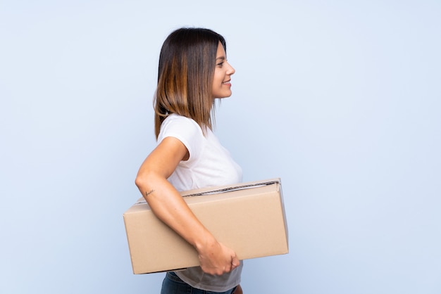 Молодая женщина над изолированным синим держит коробку, чтобы переместить ее на другое место в боковом положении