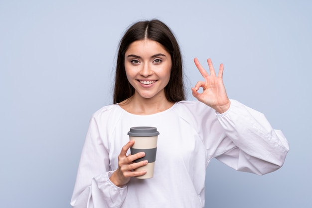 Young woman over isolated blue holding coffee to take away while making OK sign