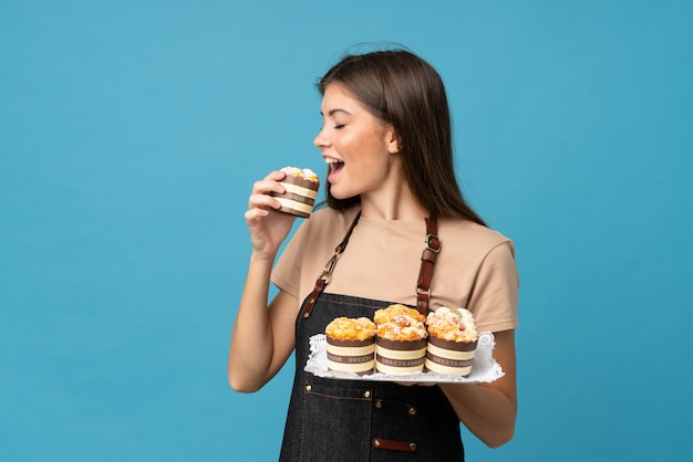 Young woman over isolated blue holding mini cakes and eating it