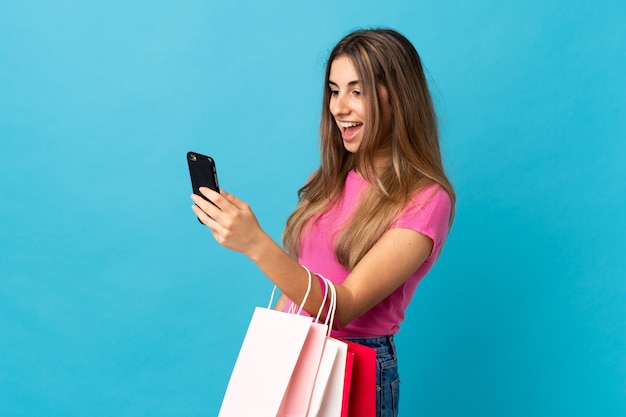 Young woman on isolated blue holding shopping bags and writing a message with her cell phone to a friend