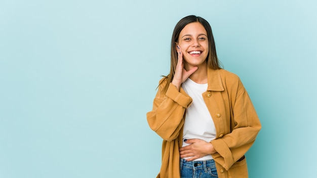 Young woman isolated on blue laughs happily and has fun keeping hands on stomach.