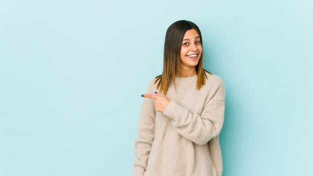 Young woman isolated on blue smiling and pointing aside, showing something at blank space.