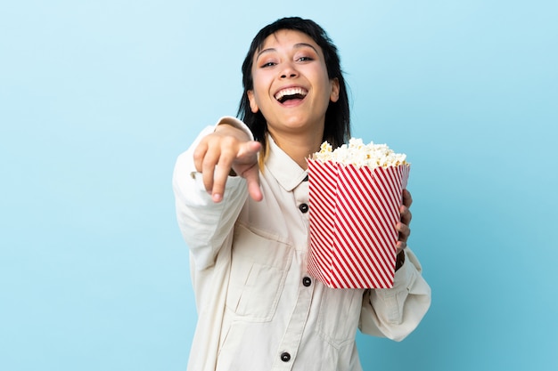 Молодая женщина над изолированным синим пространством, держащим большое ведро попкорнов, указывая спереди