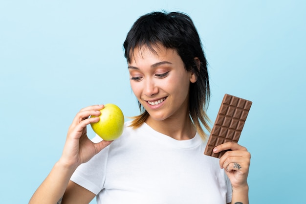 Young woman over isolated blue taking a chocolate tablet in one hand and an apple in the other