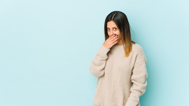 Young woman isolated on blue wall covering mouth with hands looking worried.