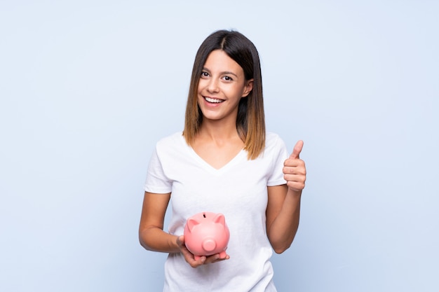 Young woman over isolated blue wall holding a big piggybank