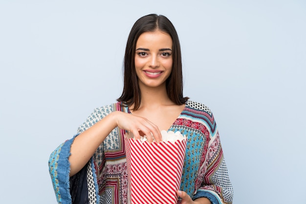 Young woman over isolated blue wall holding a bowl of popcorns