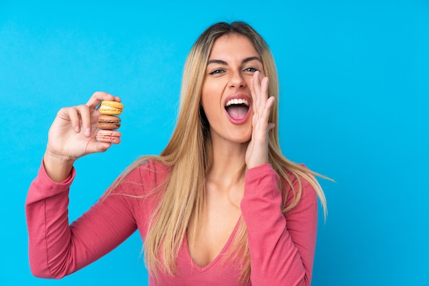 Молодая женщина над изолированной синей стеной держит красочные французские macarons и кричит