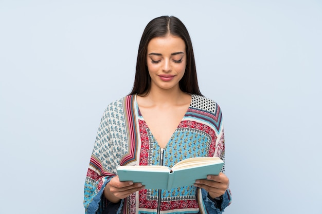 Young woman over isolated blue wall holding and reading a book