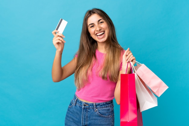 Young woman over isolated blue wall holding shopping bags and a credit card