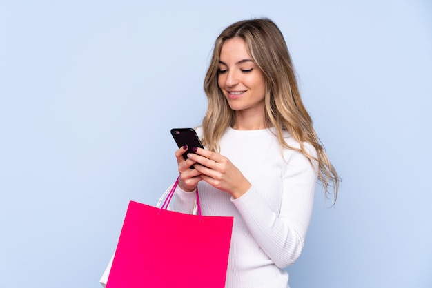 Young woman over isolated blue wall holding shopping bags and writing a message with her cell phone to a friend