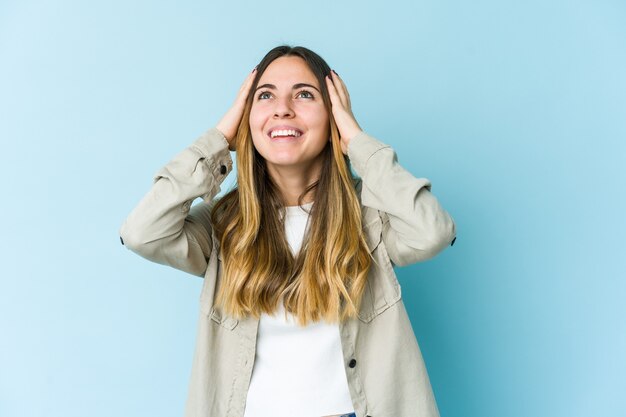 Young woman isolated on blue wall laughs joyfully keeping hands on head