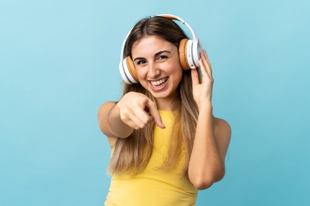 Young woman over isolated blue wall listening music and pointing to the front