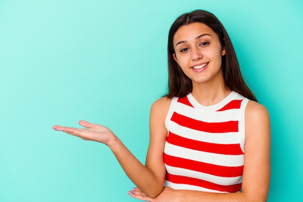 Young woman isolated on blue wall showing a copy space on a palm and holding another hand on waist