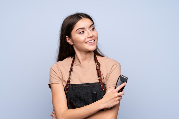 Young woman over isolated blue wall with hairdresser or barber dress and holding hair cutting machine