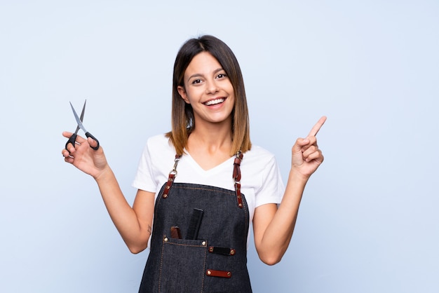 Young woman over isolated blue with hairdresser or barber dress and pointing side