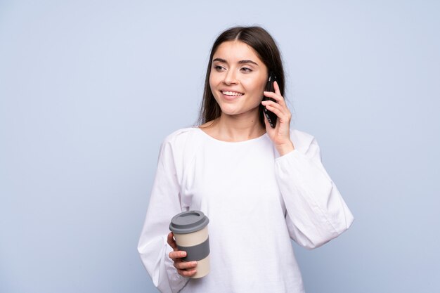 Young woman over isolated  holding coffee to take away and a mobile