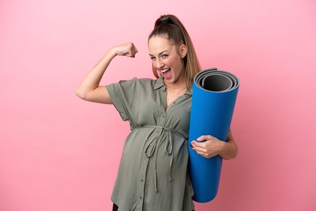 Young woman isolated on pink background pregnant and doing strong gesture while going to yoga classes