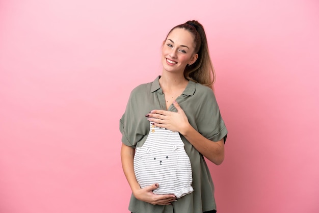 Young woman isolated on pink background pregnant and holding baby clothes