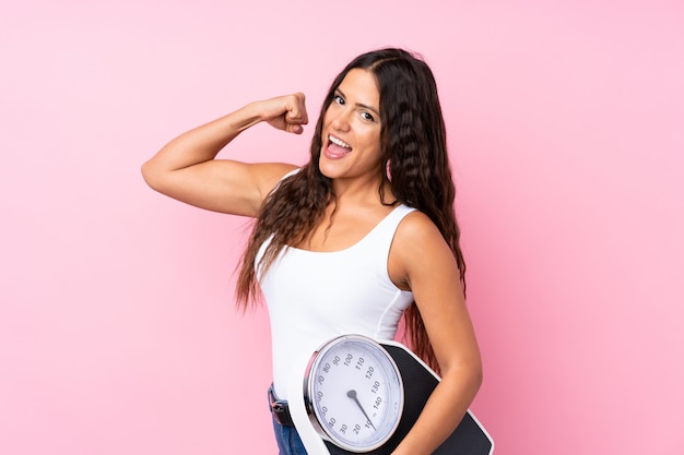 Young woman over isolated pink holding a weighing machine and doing strong gesture