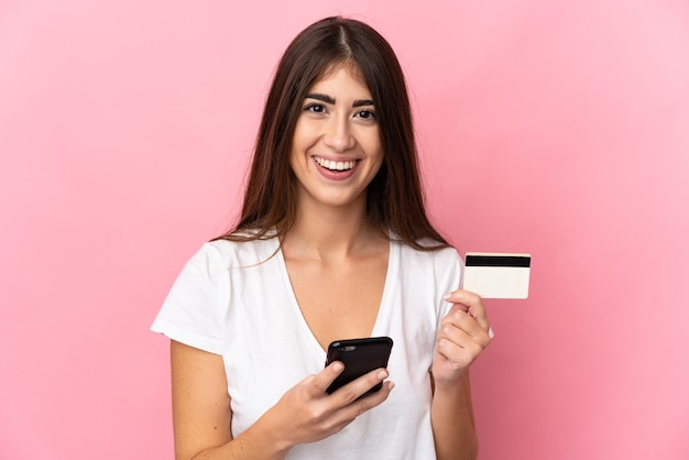 Young woman isolated on pink wall buying with the mobile and holding a credit card with surprised expression