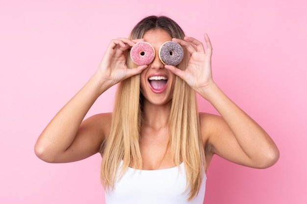 Young woman over isolated pink wall holding donuts in an eye