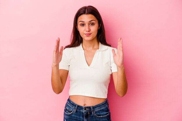 Young woman isolated on pink wall holding something little with forefingers, smiling and confident