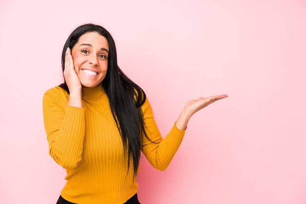 Young woman isolated on a pink wall holds copy space on a palm, keep hand over cheek Amazed and delighted.