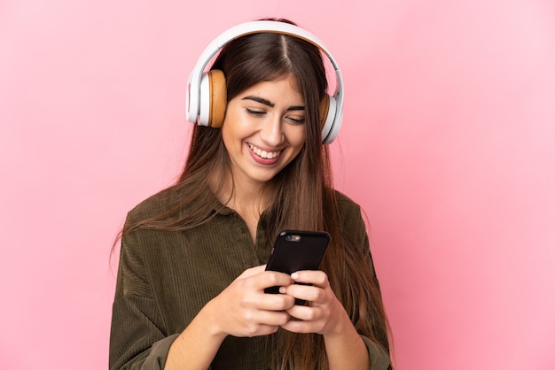 Молодая женщина изолирована на розовой стене, слушает музыку и смотрит на мобильный
