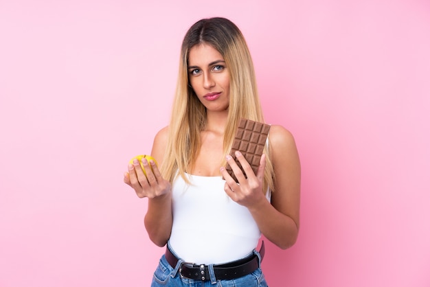 Foto giovane donna sopra la parete rosa isolata che prende una compressa di cioccolato in una mano e una mela nell'altra