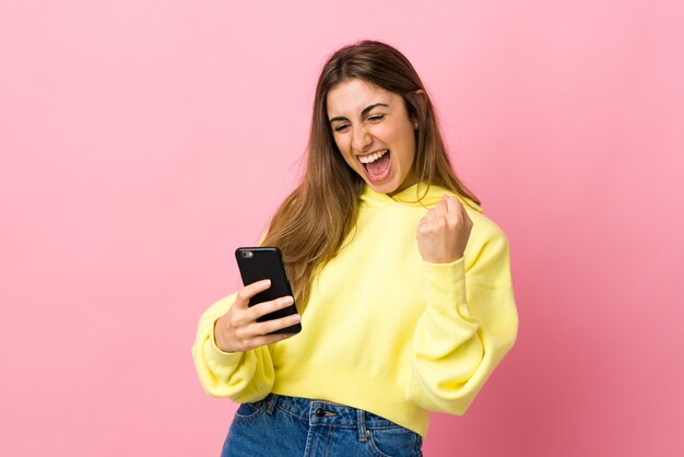 Young woman over isolated pink wall with phone in victory position